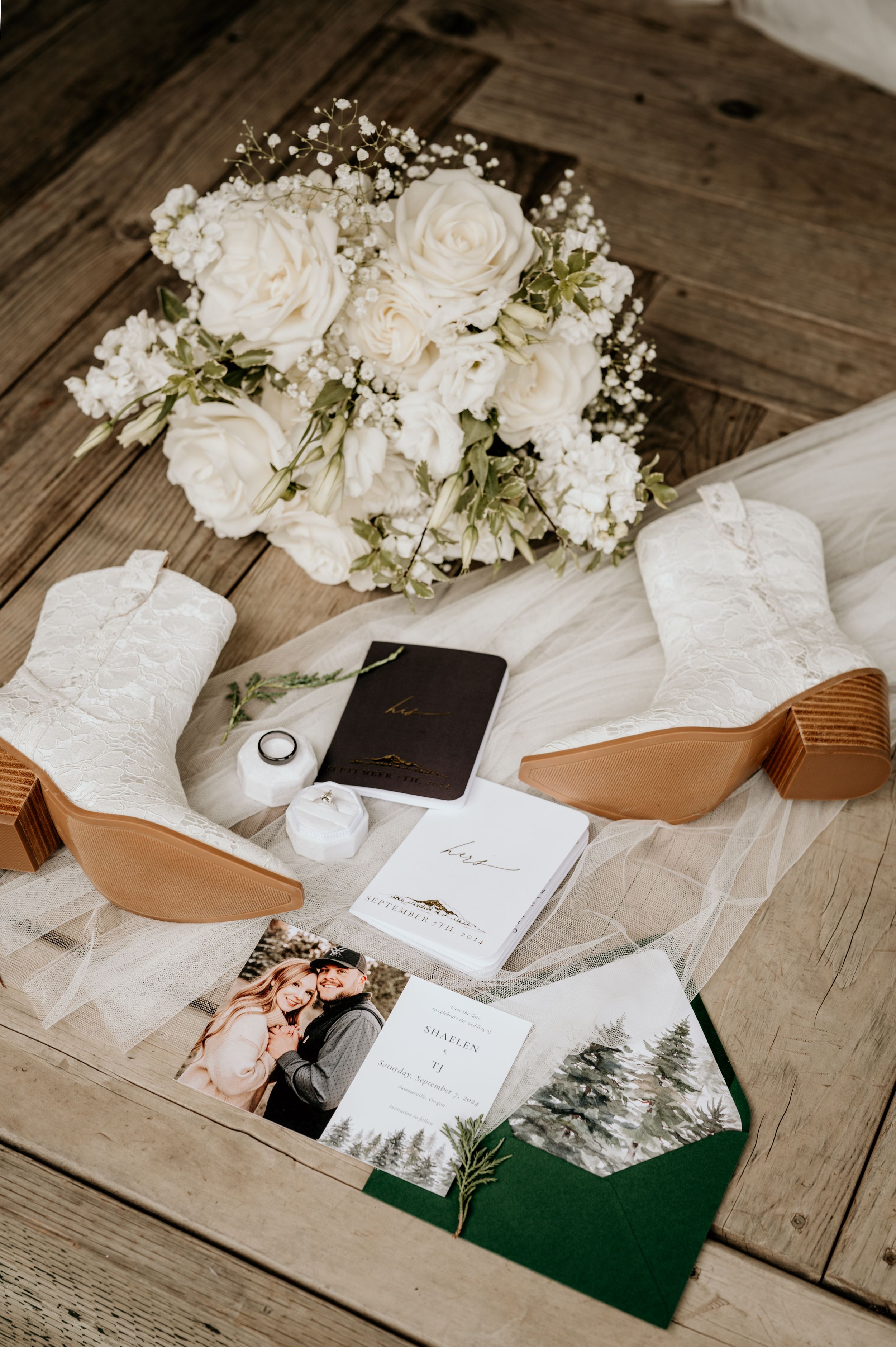 Flaylay of wedding details arranged on a wooden surface including a white flower bouquet, lace-patterned white cowboy boots, and rings.