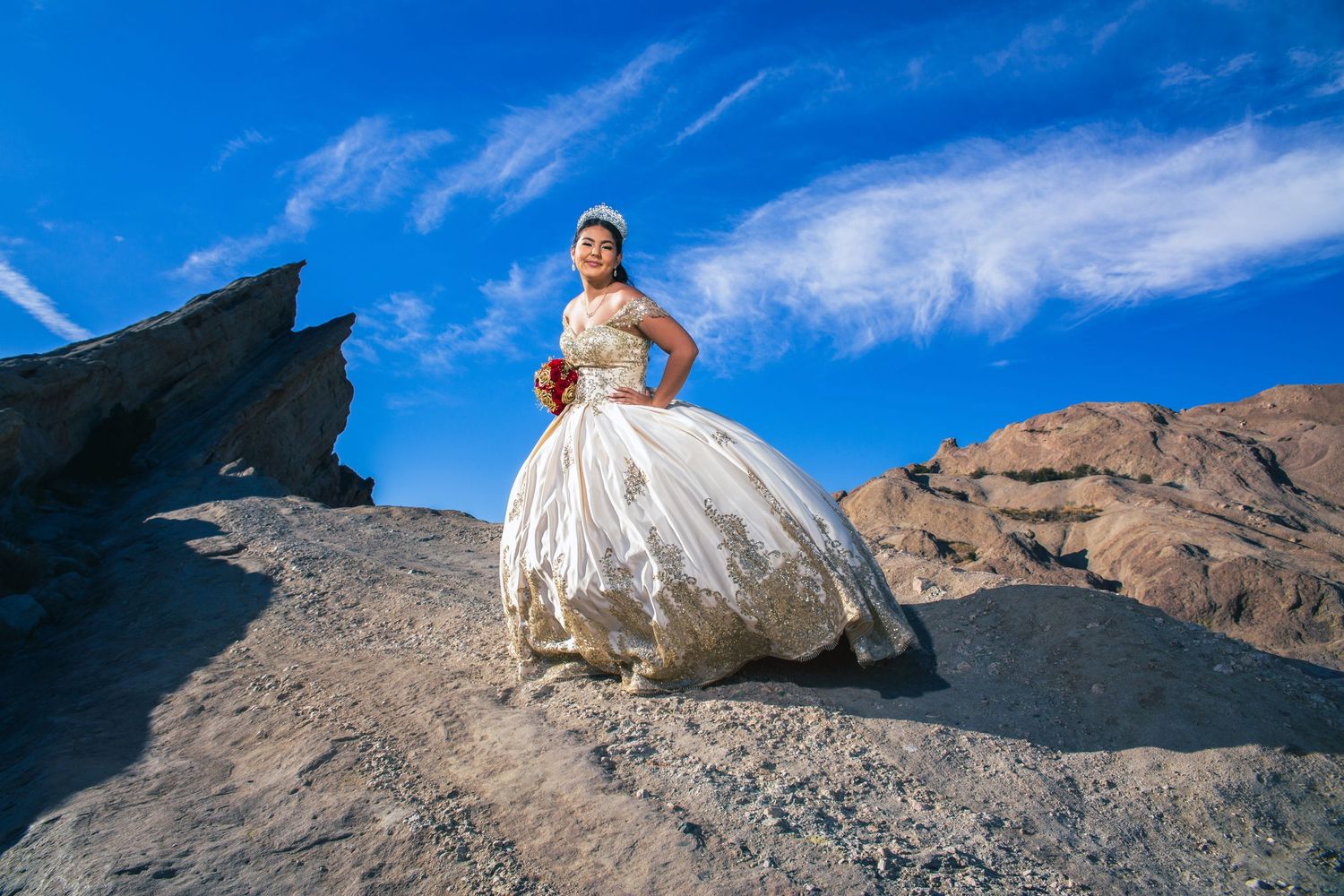 A bride in a voluminous white ball gown stands on dramatic rocky terrain against a vivid blue desert sky.