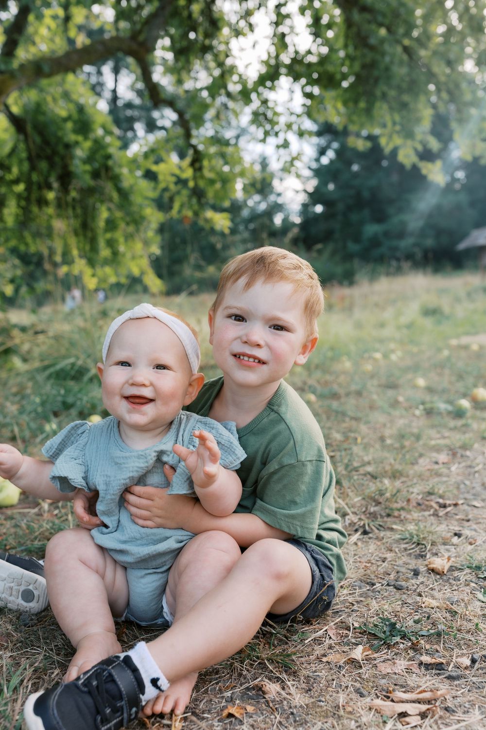 Smiling red-haired boy holding a laughing baby girl outdoors under a tree.