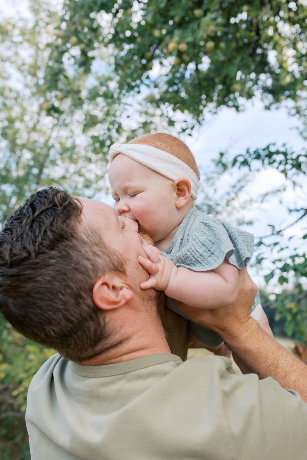 Father lifting and kissing his infant daughter wearing a white headband outdoors.