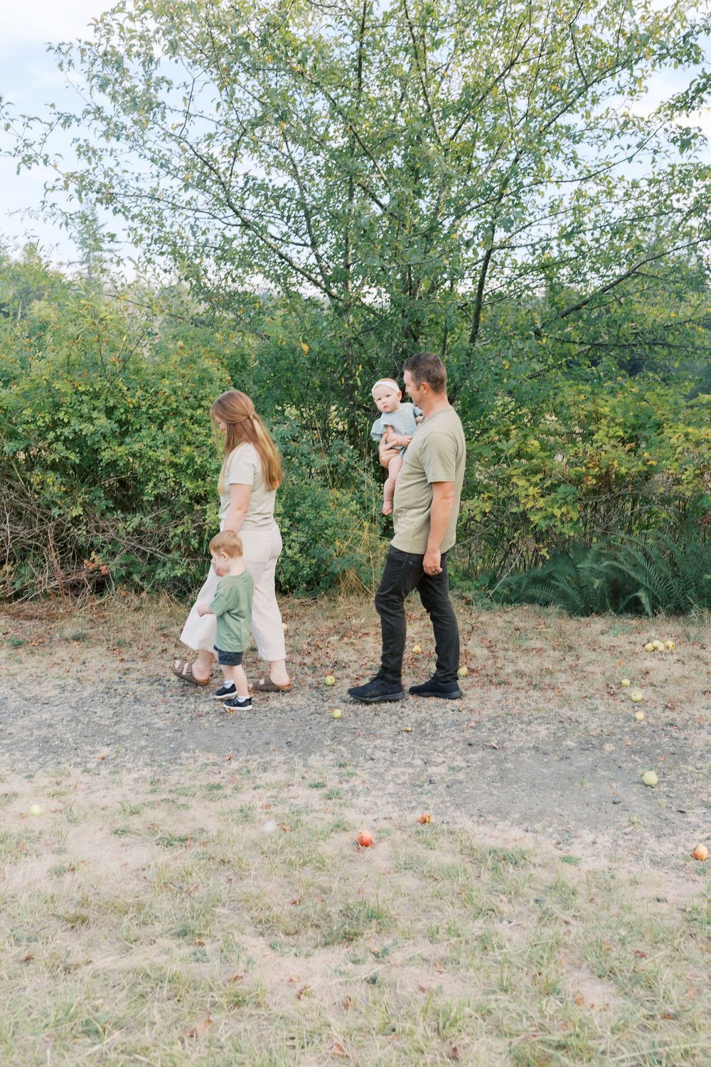 A young family with two small children walks along a dirt path surrounded by green trees and bushes outdoors.