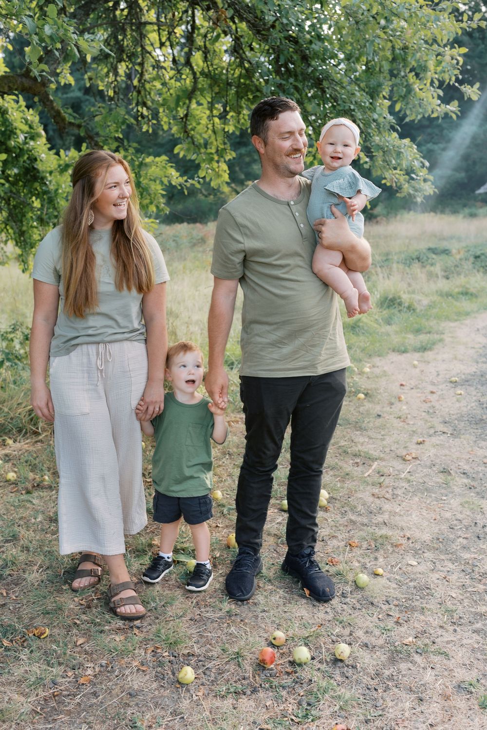 A smiling family of four stands under an apple tree, parents holding two young children in a lush green setting.