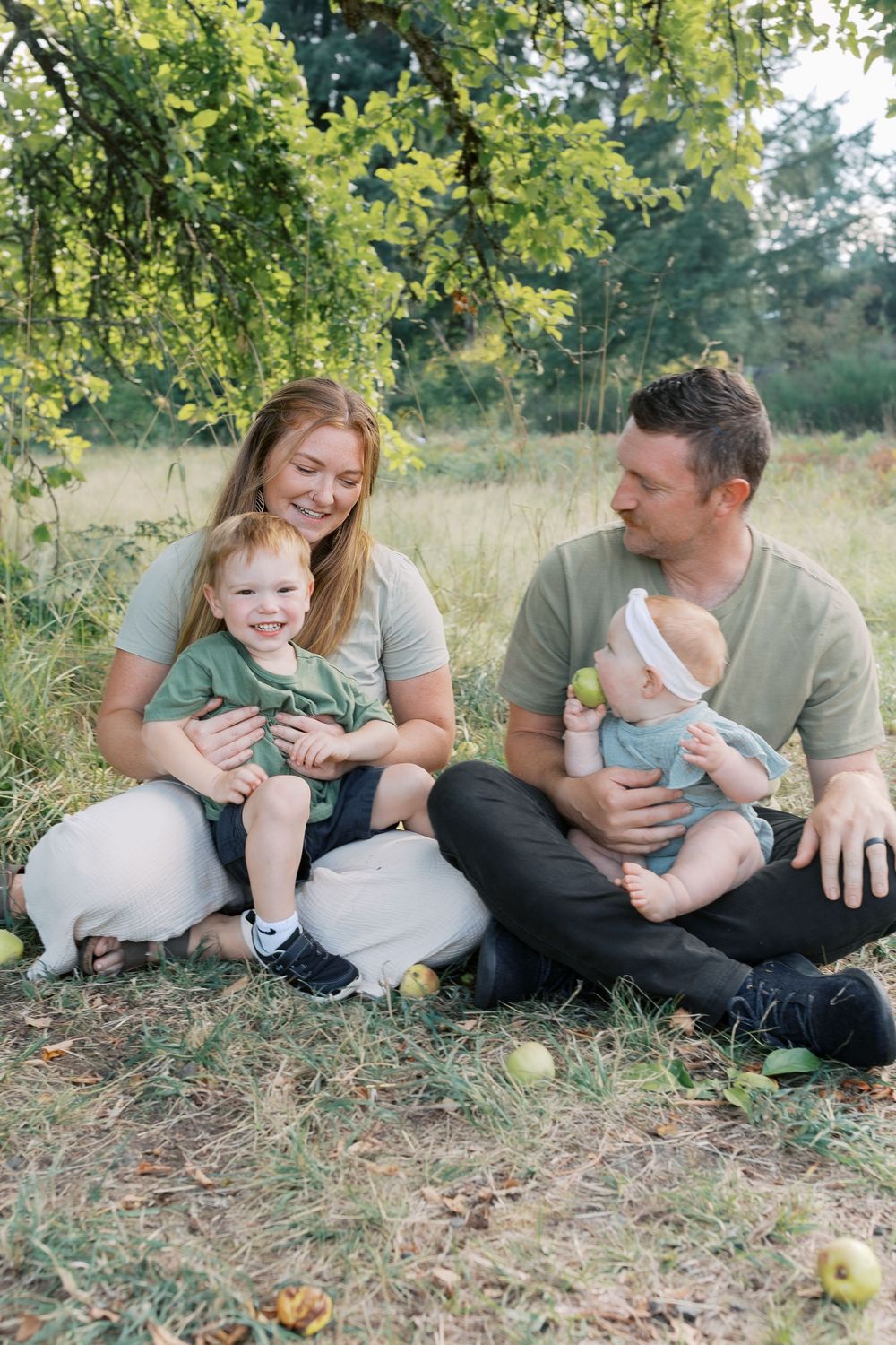 A happy family sits on grass under an apple tree, parents smiling at their toddler and baby holding a green apple.