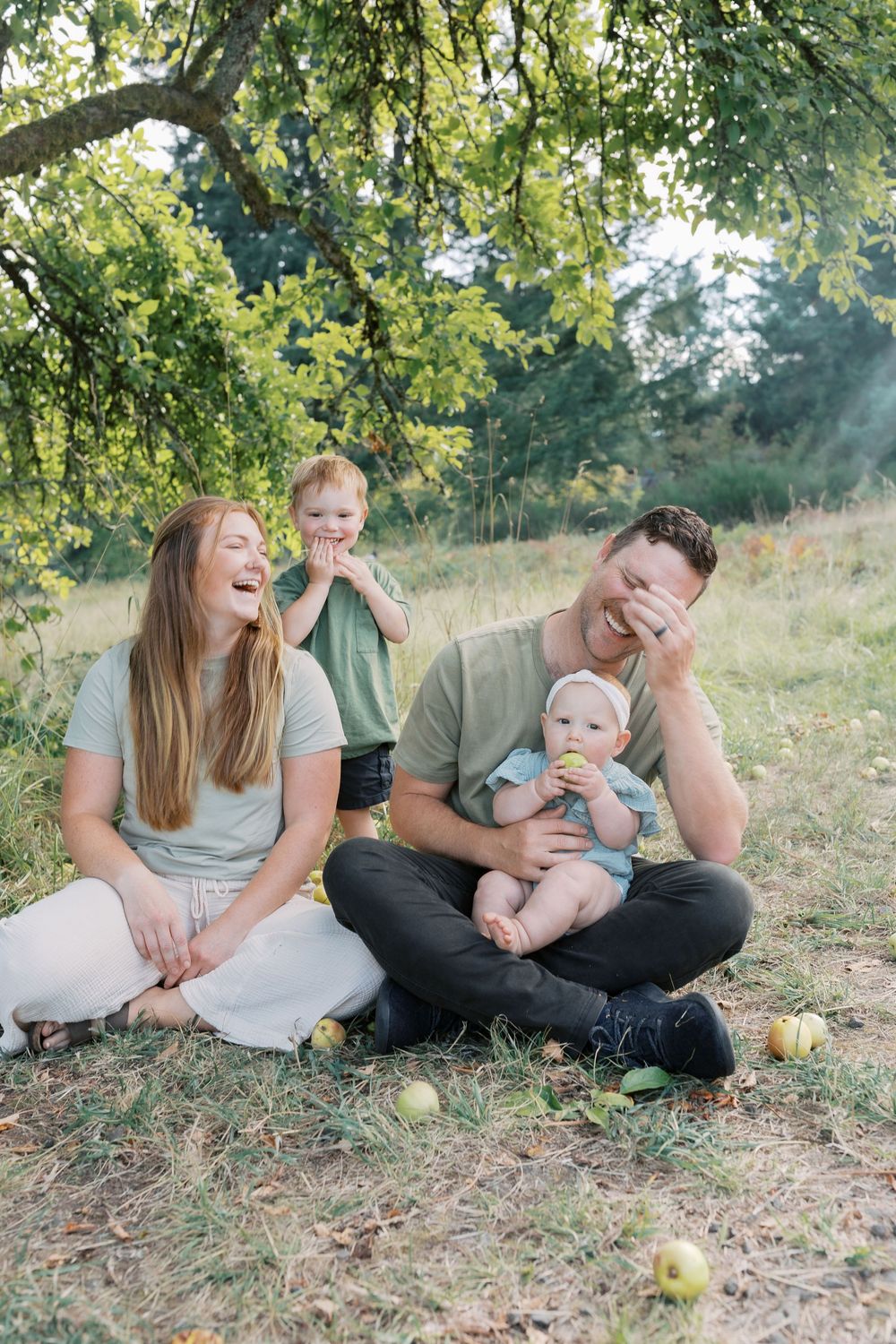 A joyful family laughs together while sitting on grass under an apple tree with fallen apples around them.