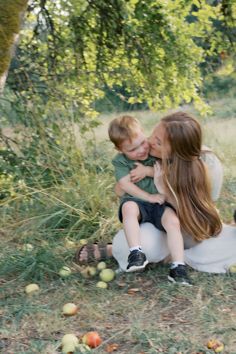 A mother crouches down to hug and kiss her smiling toddler boy near an apple tree in a grassy outdoor setting.