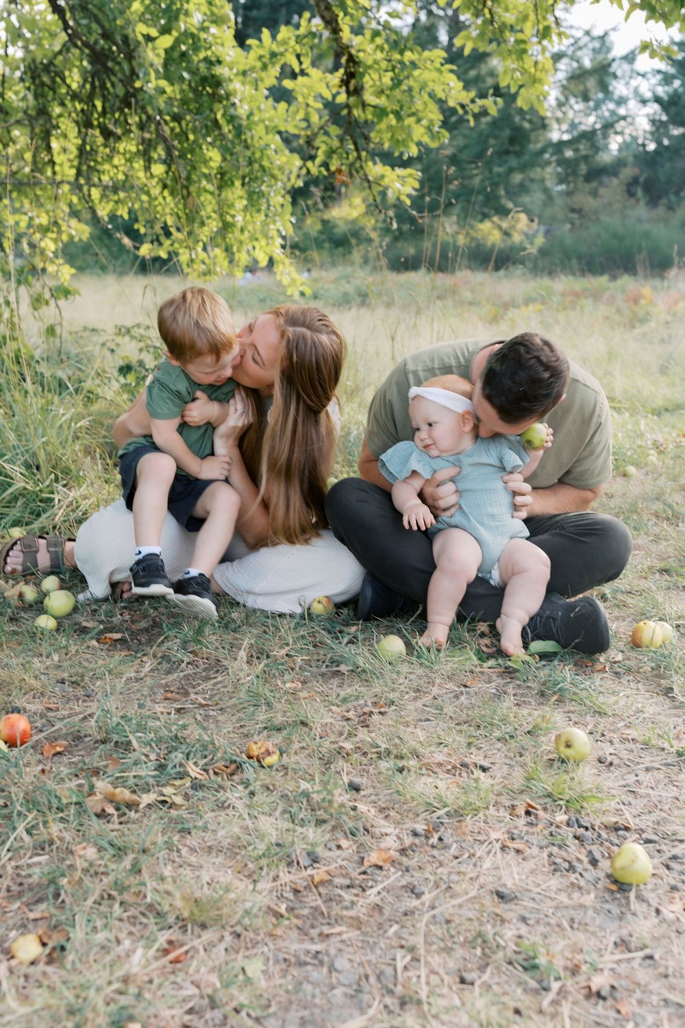 Parents lovingly kiss their two young children while sitting together on grass beneath a leafy apple tree outdoors.