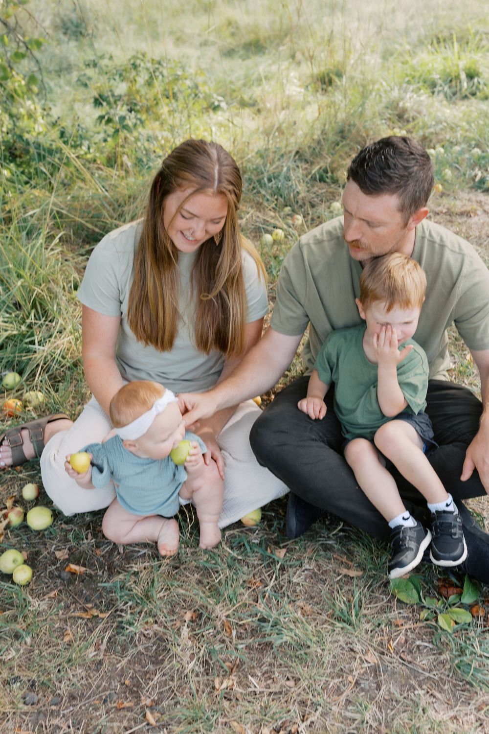 A family of four sits on grass under an apple tree as parents watch their baby and toddler play with apples.