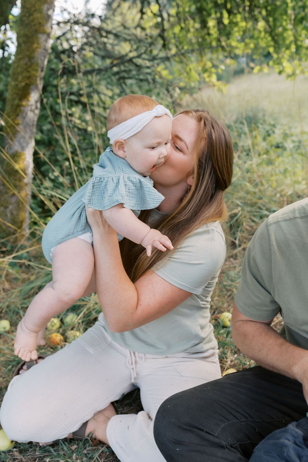 A mother lifts and kisses her smiling baby girl wearing a white headband while sitting outdoors near an apple tree.