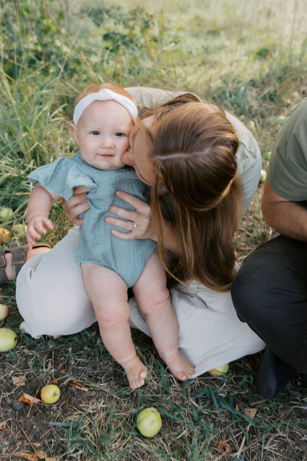 A mother gently kisses her baby girl in a blue romper and white headband while sitting on grass near apple trees.