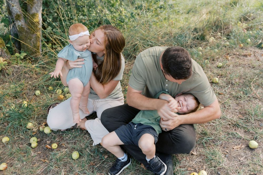 A playful family sits on grass as a mother kisses her baby while the father playfully wrestles with their toddler.