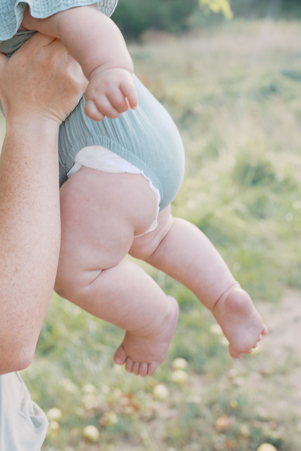 Adult hands holding a chubby baby wearing a light blue diaper outdoors in a green field.