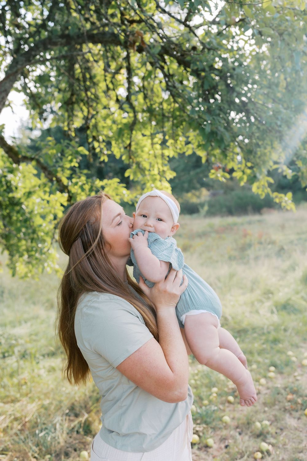 A mother kisses her smiling baby outdoors under a green tree in warm sunlight.