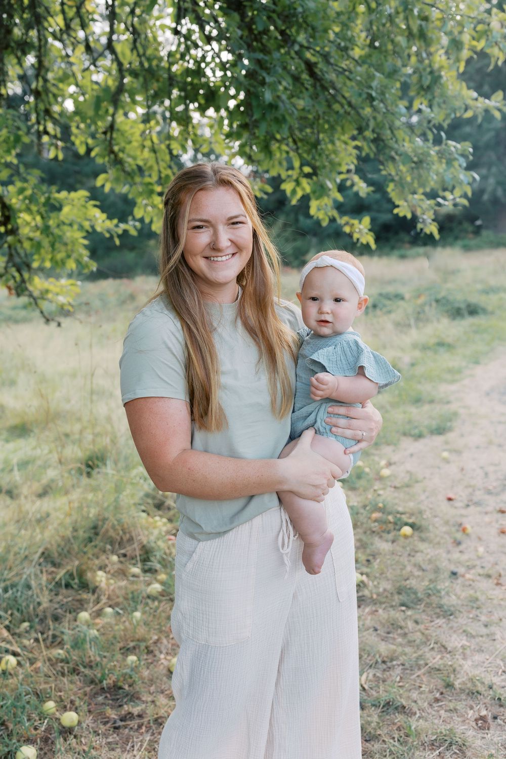 Smiling young woman in sage green top holding an infant in a white headband outdoors in a lush green park.