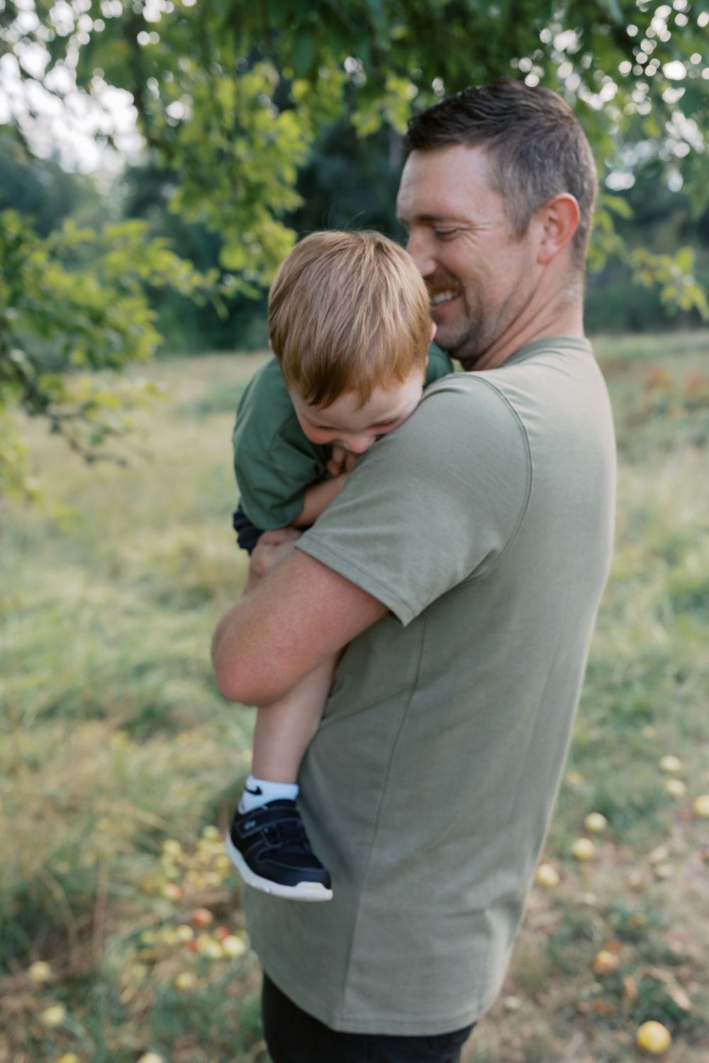 A smiling man in olive green t-shirt holds a young blonde boy outdoors in a lush green orchard setting.