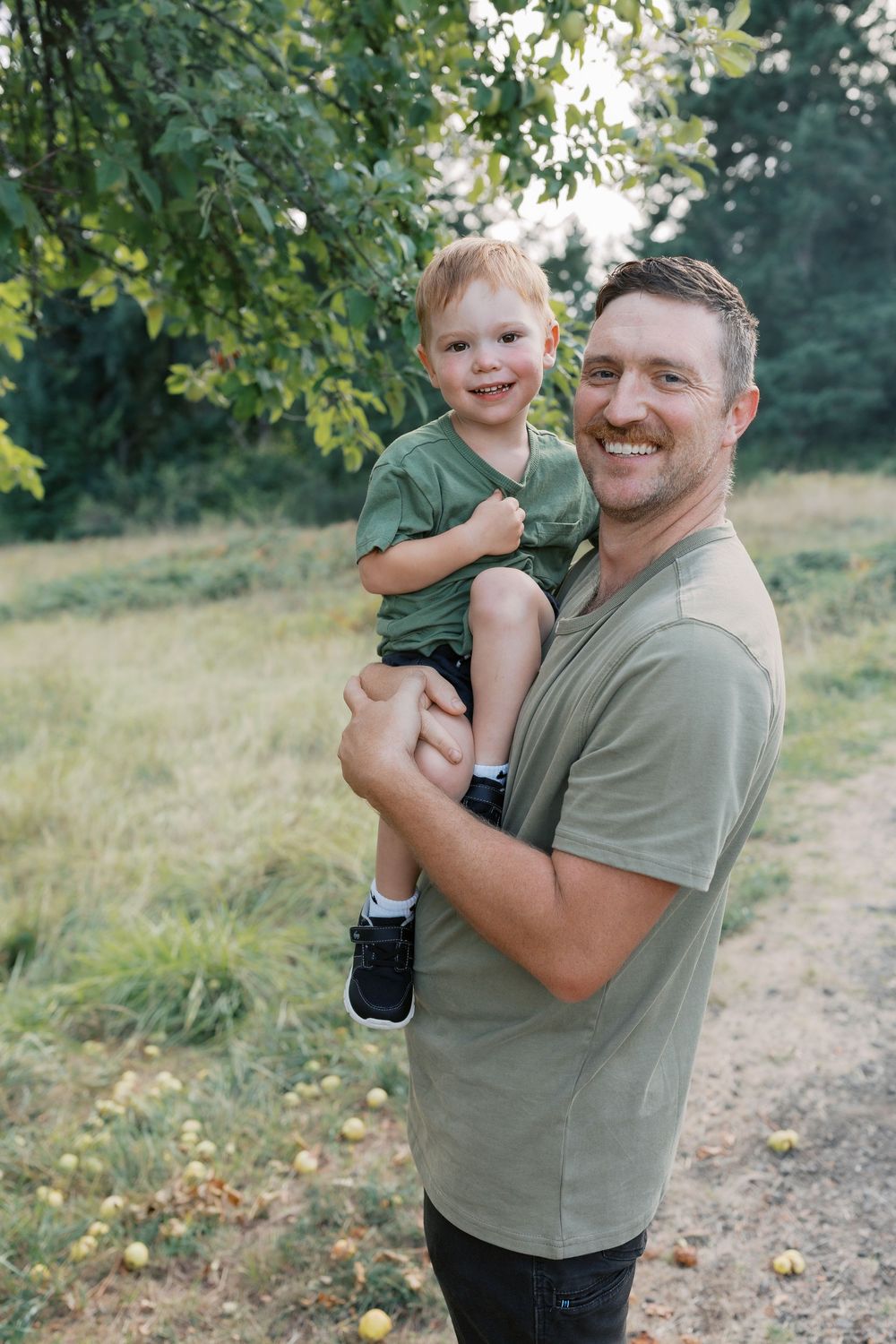 A smiling man in a green shirt holds a happy toddler boy outdoors near apple trees in a lush green park.