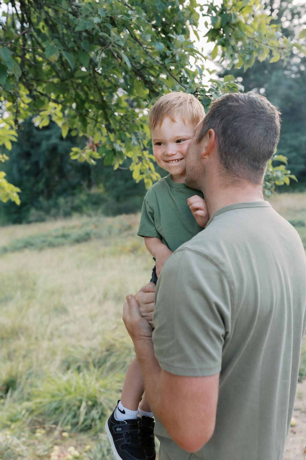 A smiling young boy in a green shirt being held by his father outdoors near green trees.