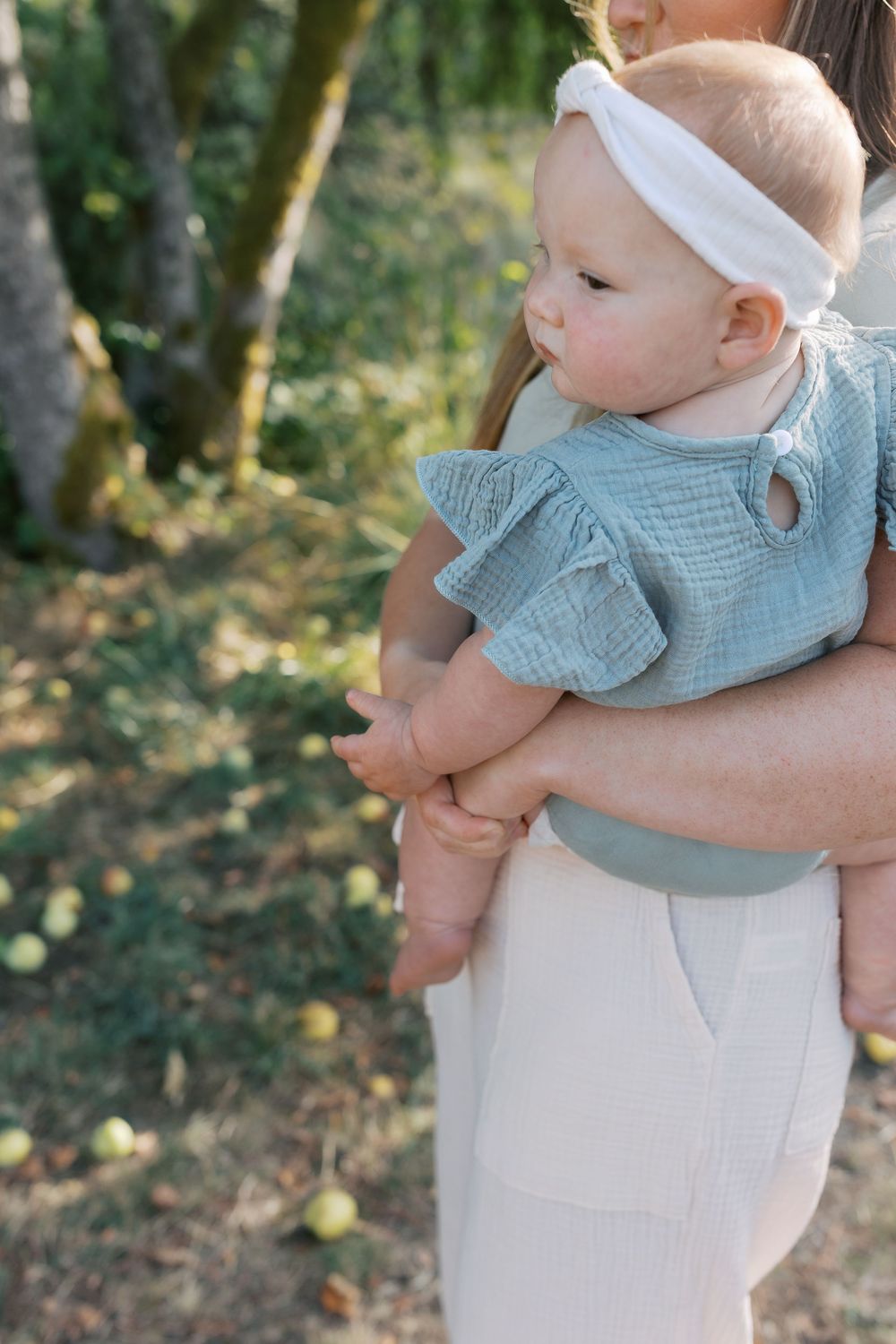 A baby in a blue ruffle top and white headband being held outdoors near apple trees.