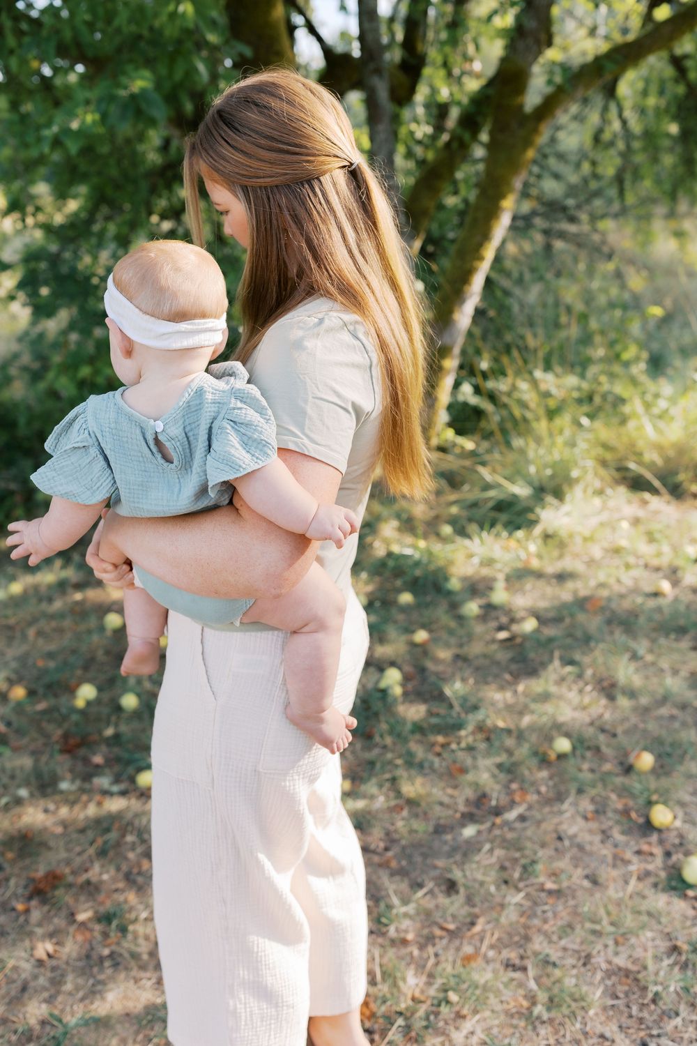 Young mother holding baby girl in white headband outdoors near apple tree in summer garden.