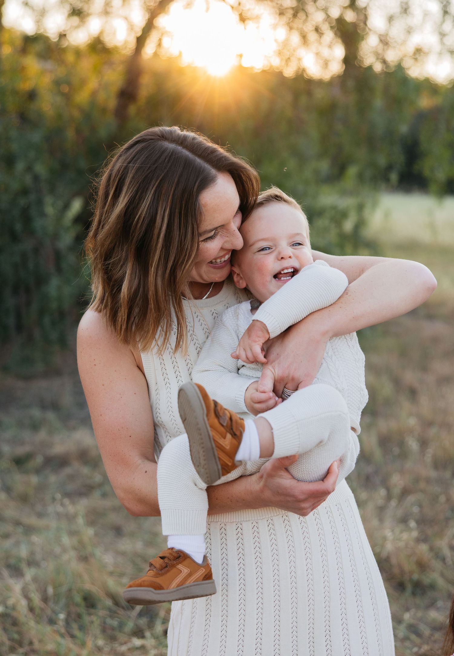 Mother and baby share tender moment in sunset field wearing white knit dress.