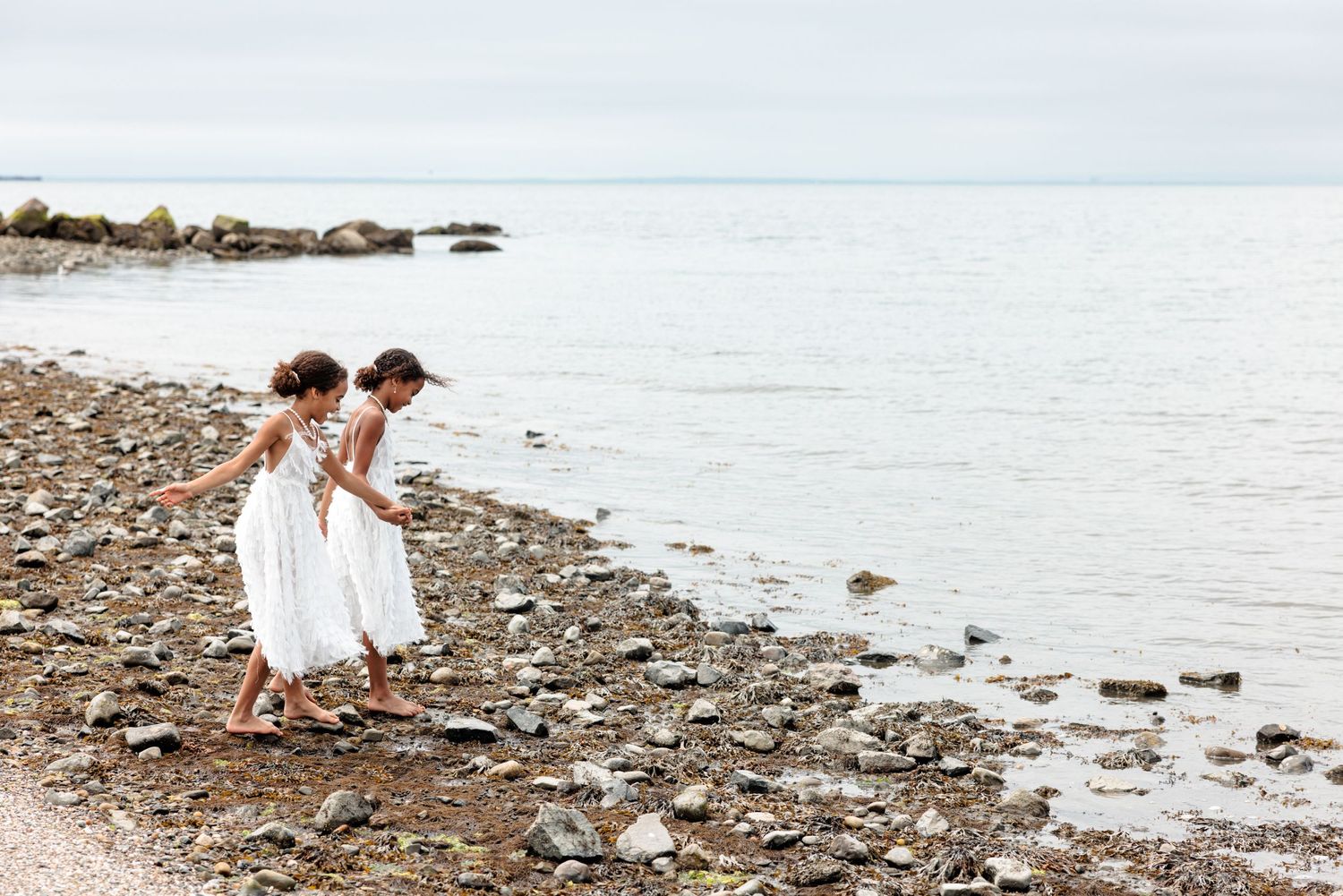 Two children in white dresses explore a rocky beach shoreline together.