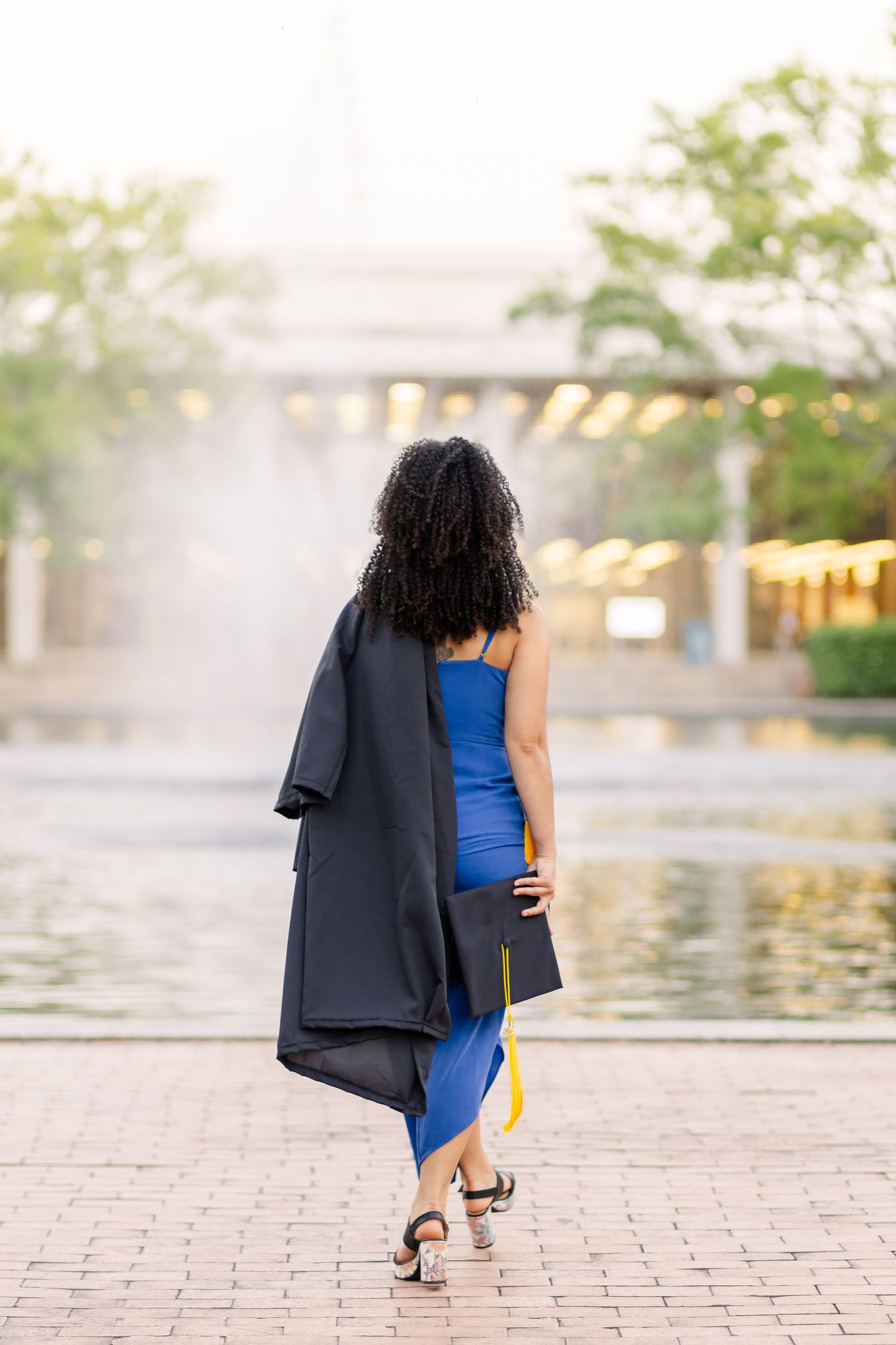 A woman in a blue dress and black coat walks near a fountain at sunset.