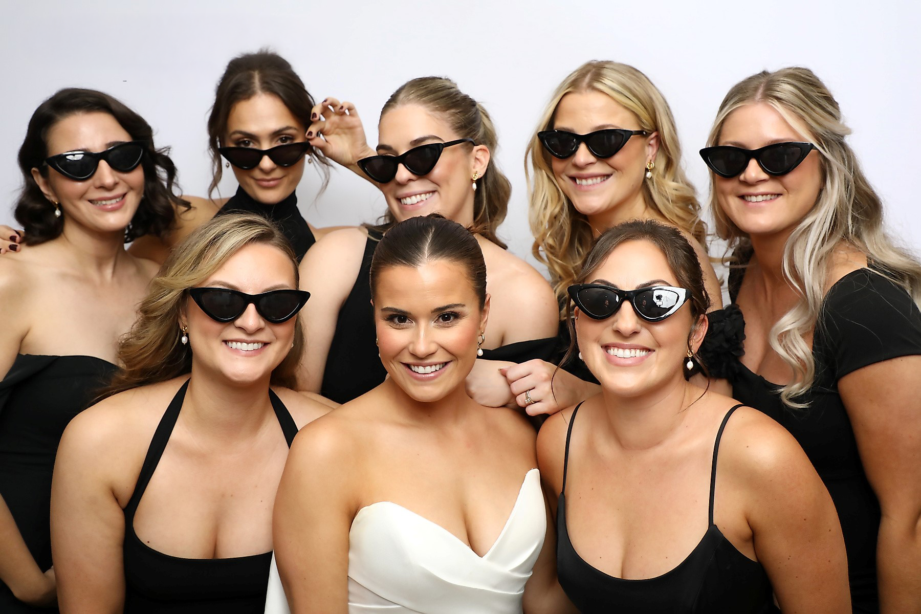 A group wearing black dresses and sunglasses poses together with someone in a white dress at the center.
