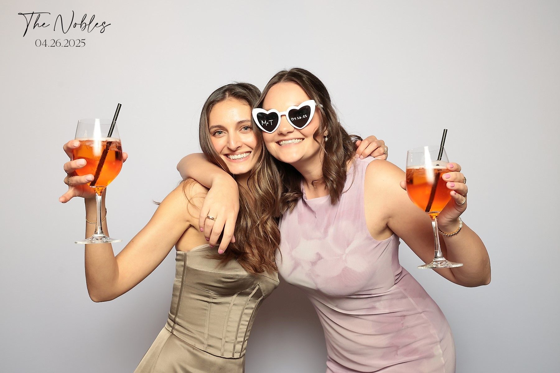 Two friends in fancy dresses hold up orange cocktails while posing together against a light gray background.