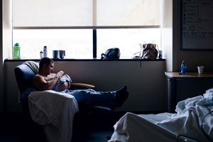 Father holding newborn baby in hospital room chair beside window, tender bonding moment in dimly lit maternity ward.