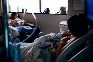 New parents rest in a hospital room, with mother breastfeeding newborn baby while father sits nearby.