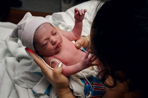 Newborn baby held by mother in hospital shortly after birth, wearing pink hat with umbilical cord clamp visible.