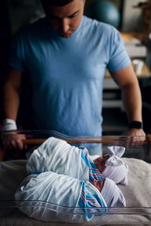 A new father in a blue shirt gazes lovingly at his swaddled newborn baby in a hospital bassinet.