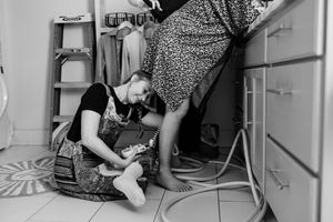 Black and white photo of a woman sitting on kitchen floor working on plumbing while another person stands nearby.