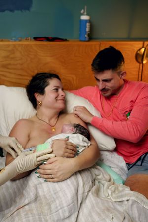 A new mother holds her newborn baby in a hospital bed while the father in a pink shirt looks on lovingly.