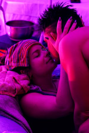 Two people sharing an intimate moment under purple lighting, one wearing a colorful head wrap while lying down.