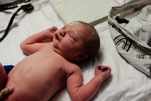 Newborn baby lying on white blanket in hospital, eyes closed, tiny fists clenched, with medical equipment nearby.