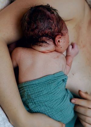 Newborn baby with dark hair resting on mother's chest, partially wrapped in a teal muslin cloth after birth.