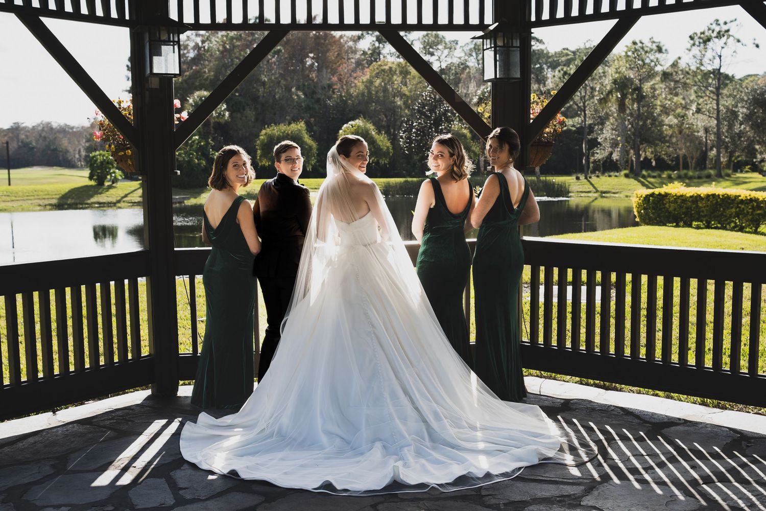 A bridal party in black gowns and a white wedding dress poses together under a gazebo on a sunny afternoon.