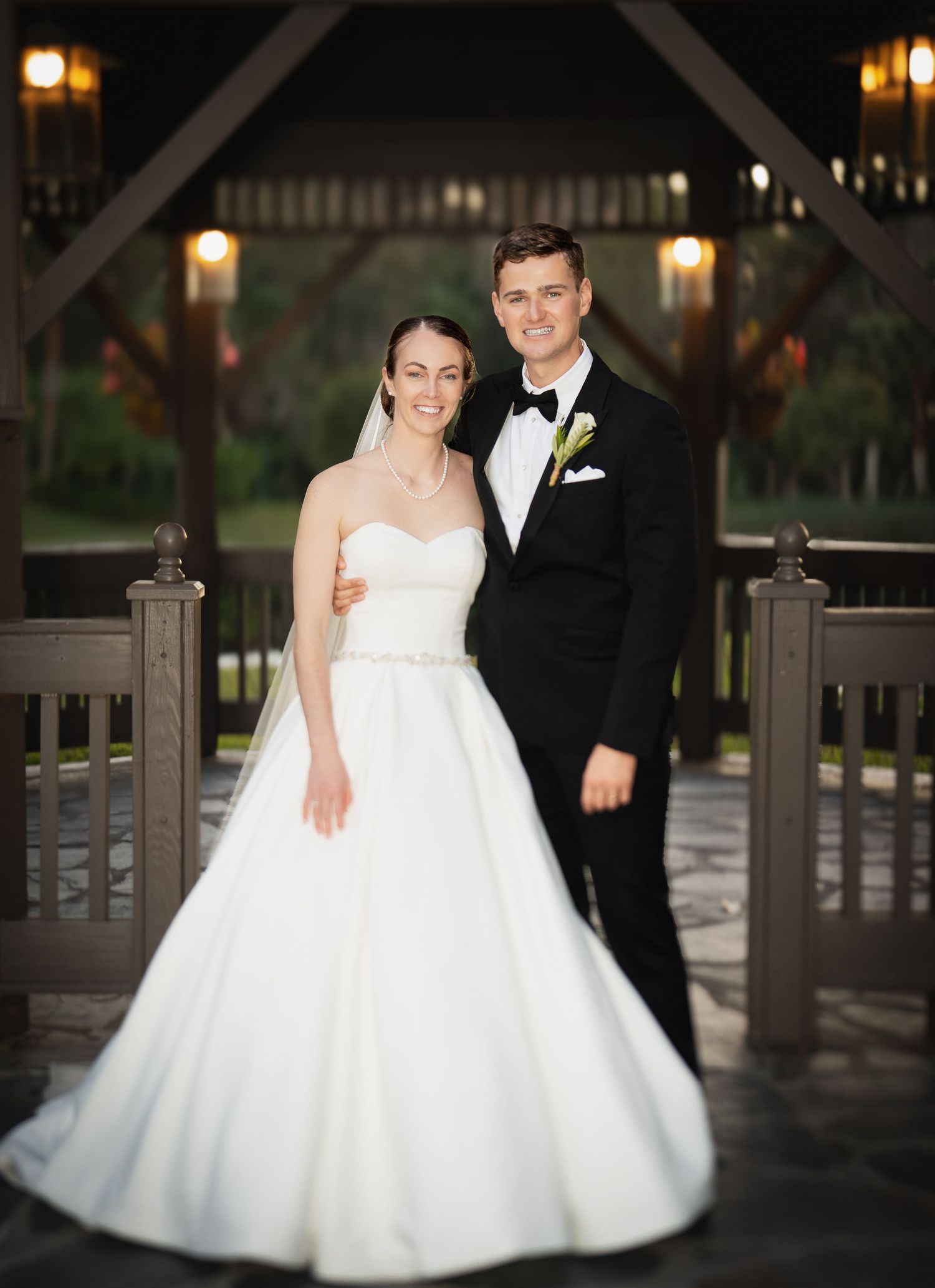 A bride and groom pose on a wooden deck at night, surrounded by glowing lanterns in elegant wedding attire.