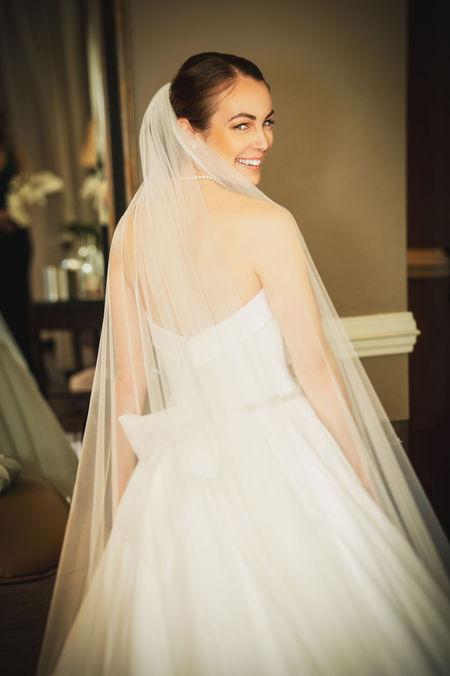 A bride in a classic white strapless wedding gown and flowing veil smiles while looking over her shoulder in warm lighting.