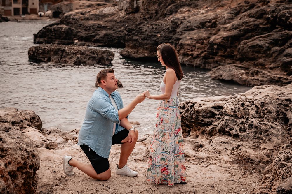 Man proposing on one knee to his girlfriend on a rocky beach during a romantic Mallorca beach proposal.