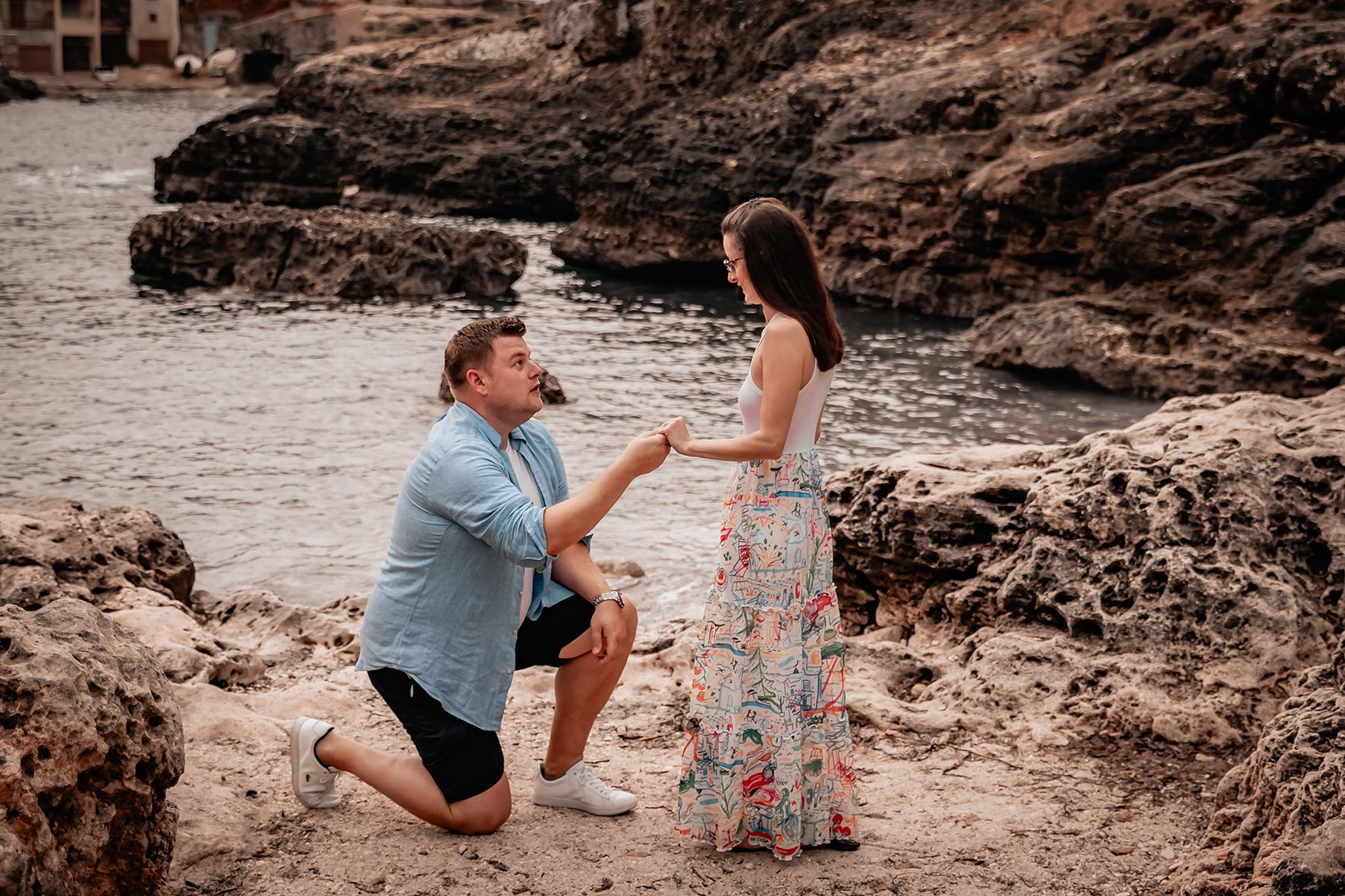 Man proposing on one knee to his girlfriend on a rocky beach during a romantic Mallorca beach proposal.