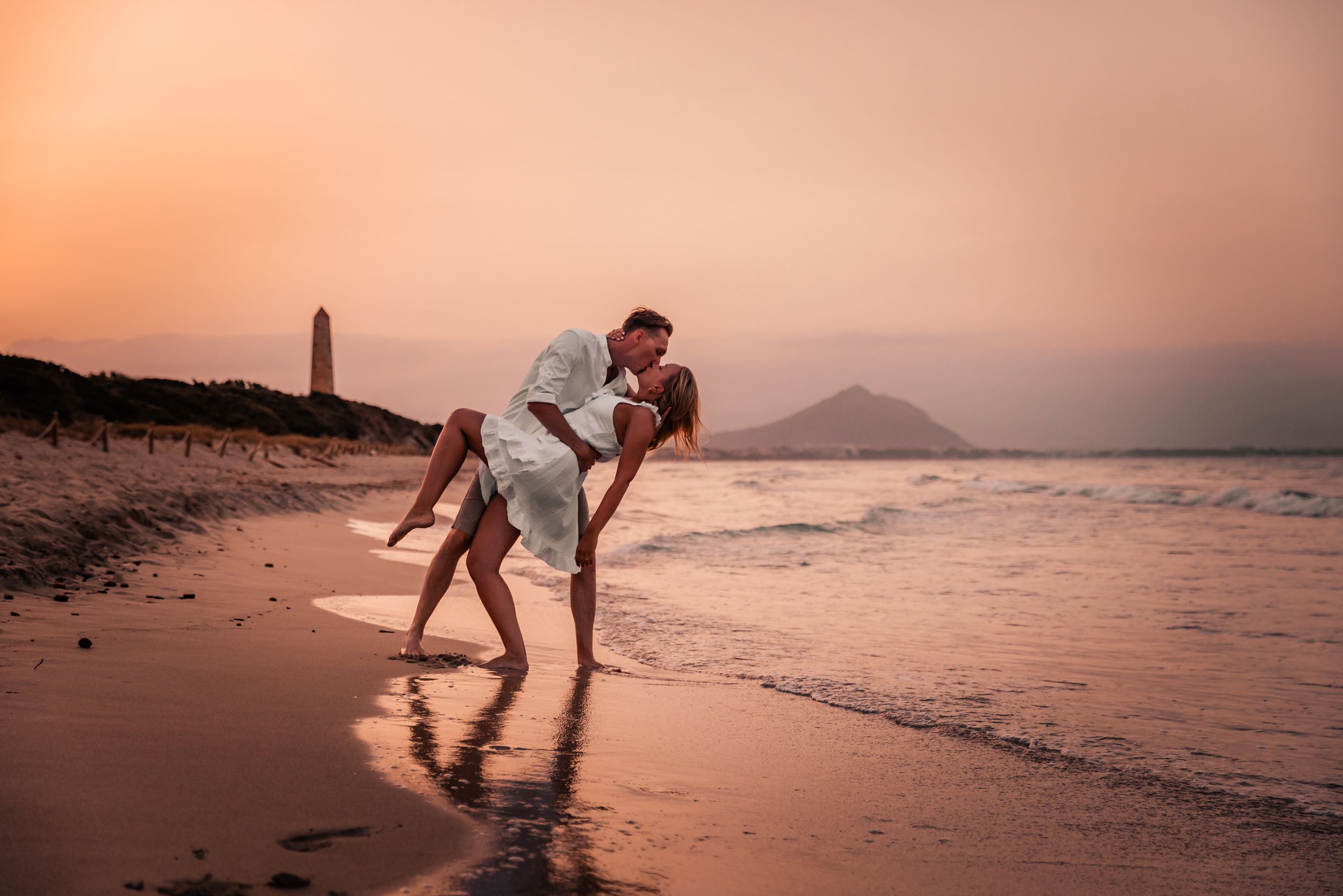 Wide shot of a couple on a sandy beach while he is holding her in a 'dip pose' while kissing.