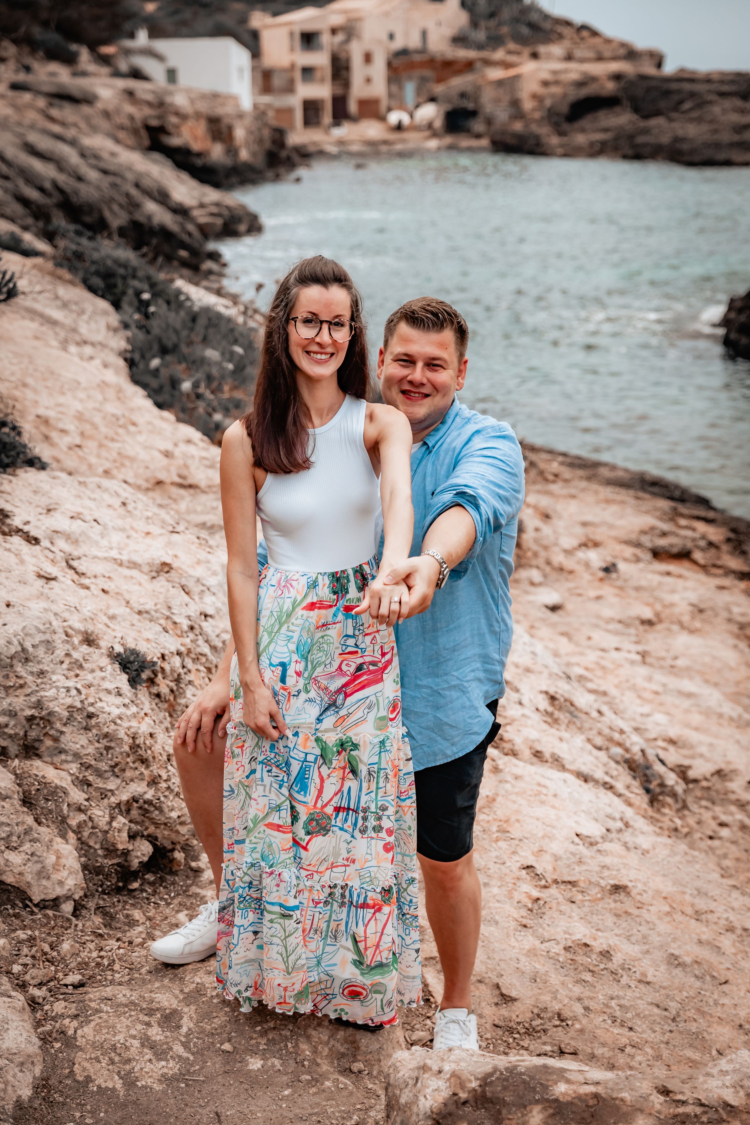 A happy proposal couple standing and smiling on a rocky beach together while she is showing her engagement ring into the camera.