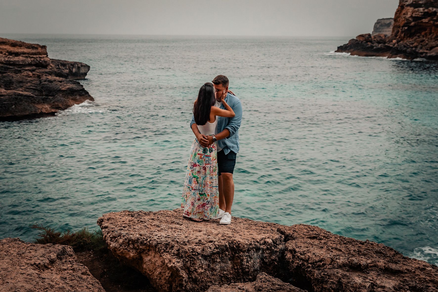 Wide view of a couple embracing and standing very close to each other on a rocky beach.
