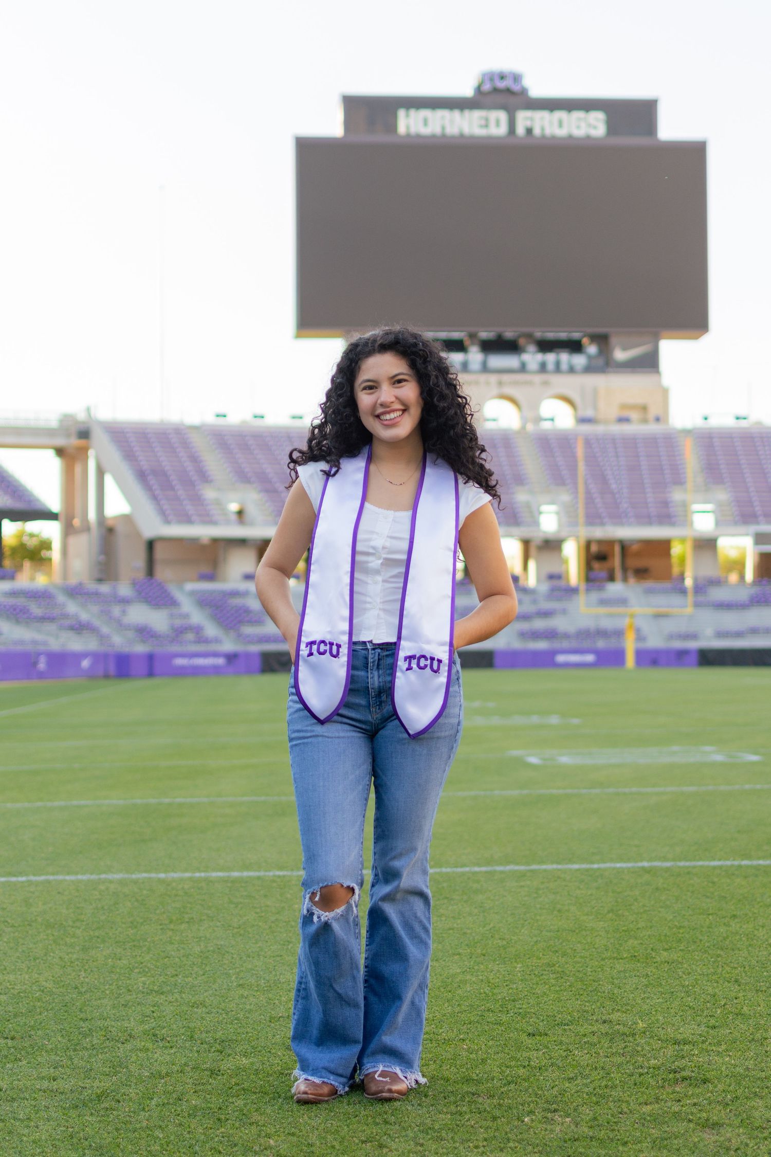 A TCU graduating senior poses with her hands in her jean pockets while wearing her white & purple grad stole, standing on the football field