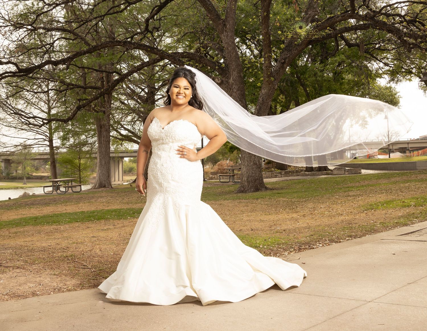Bride in fitted white mermaid gown with flowing veil posing in outdoor setting.