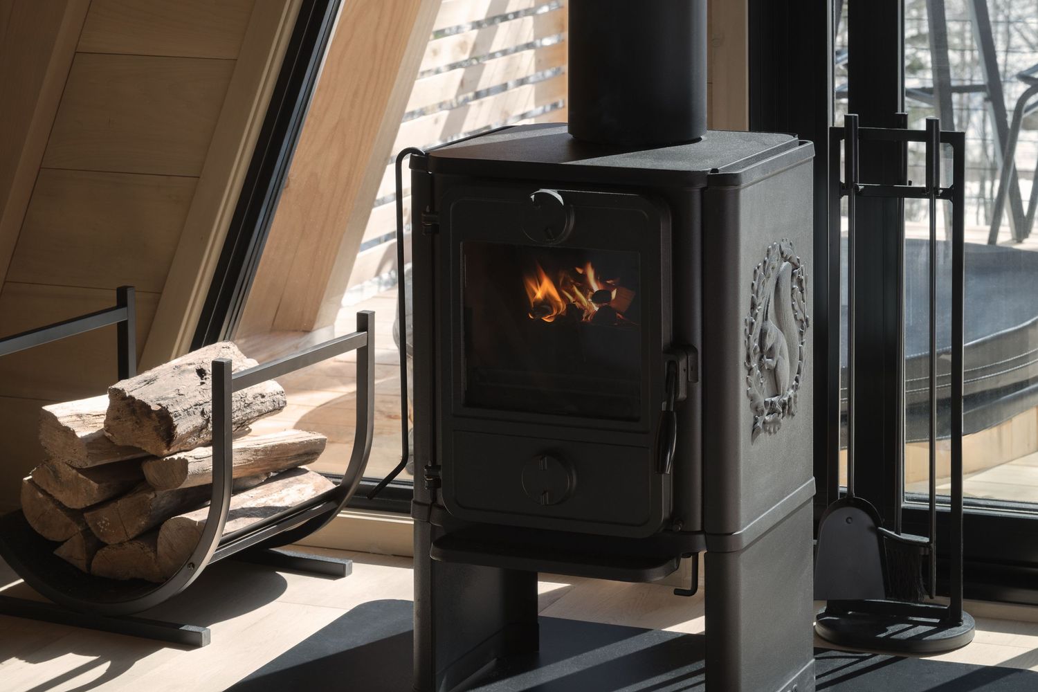 A cozy black wood-burning stove with a glowing fire inside sits next to stacked firewood in a warm sunlit room.