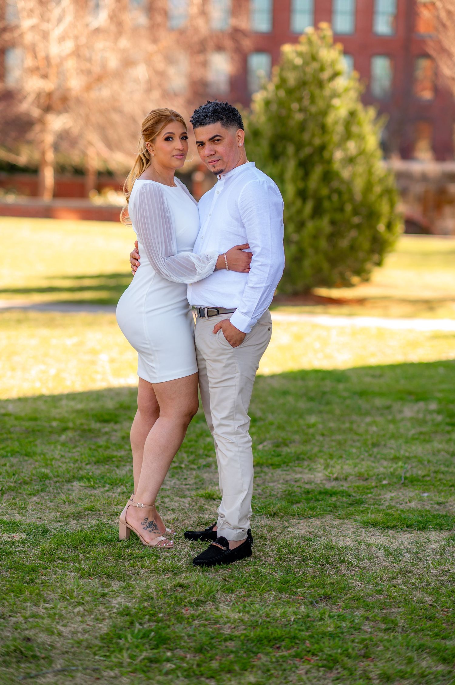 A romantic couple in coordinated white outfits posing on grass near brick building.