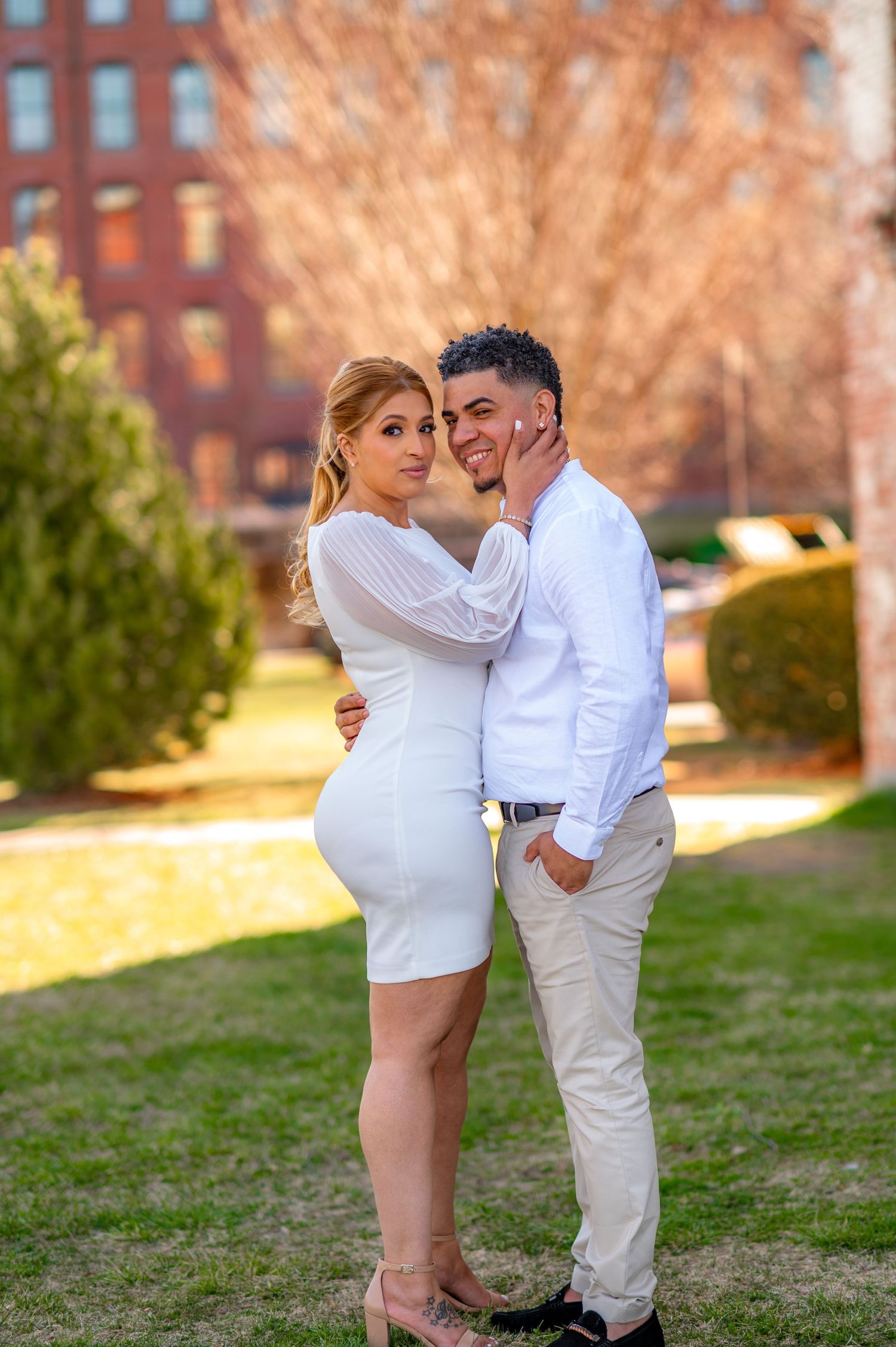 A couple in matching white outfits embracing on grass with brick building in background.