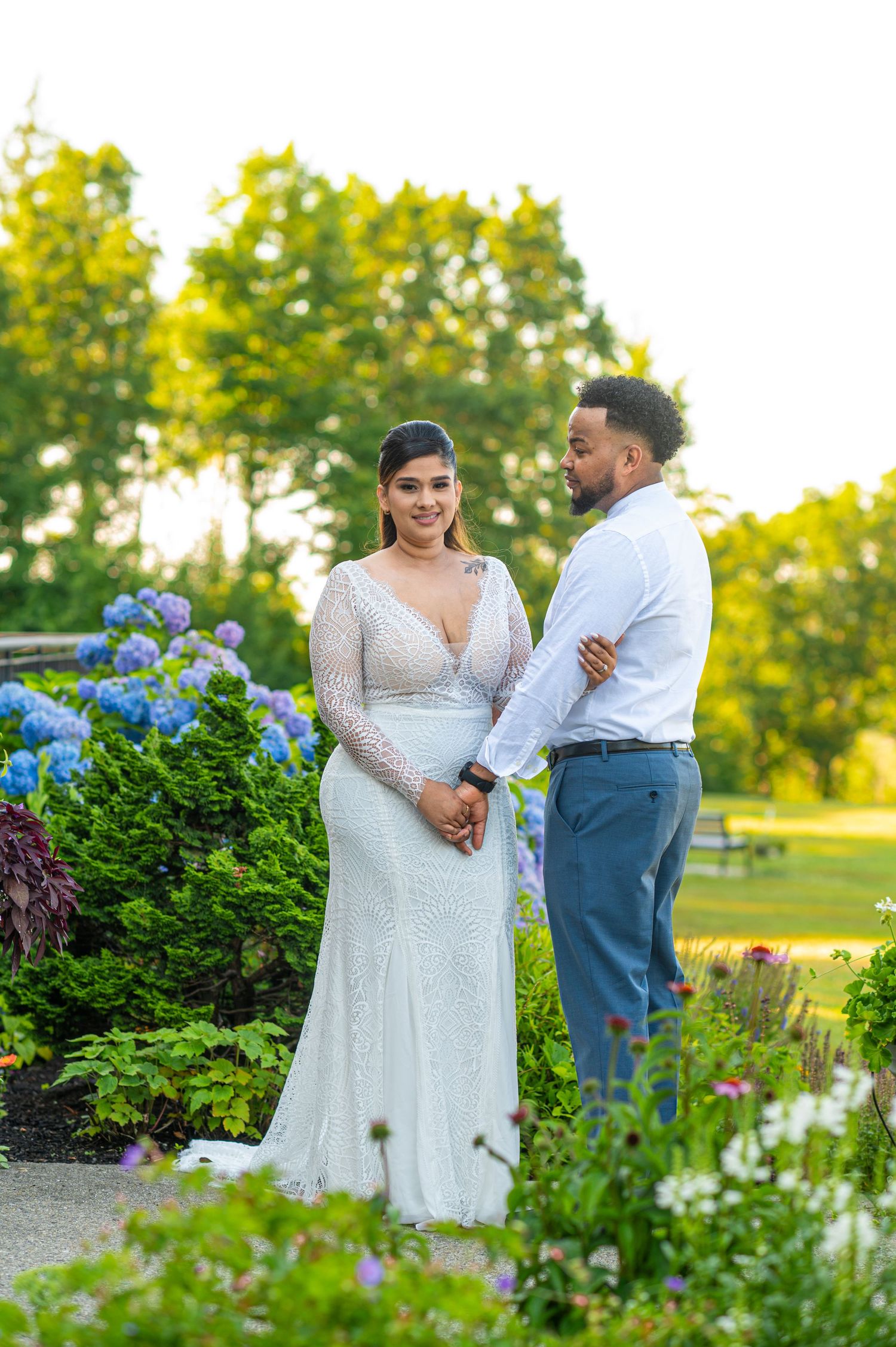Bride in lace gown and groom in blue suit hold hands in a lush garden with hydrangeas at golden hour.